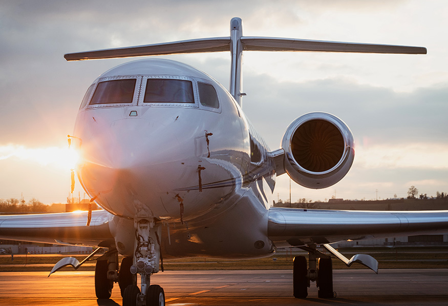 A private jet is pictured on a tarmac at sunset, with the sun gleaming behind it, highlighting its sleek design and twin engines.