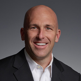 A smiling person with a shaved head is pictured against a grey background. They are wearing a dark suit jacket with a white shirt.