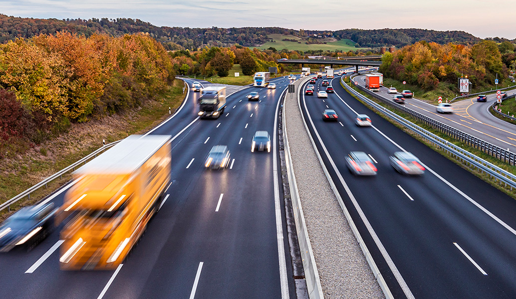 A busy multi-lane highway with cars and trucks in motion. Surrounded by colorful autumn trees at dusk, with a bridge overhead and a clear sky.