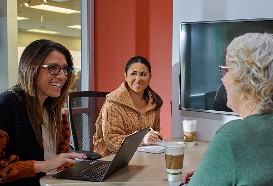 Two smiling people having a meeting with an older person, sitting at a table with a laptop and coffee cups, in a room with a red wall and TV.