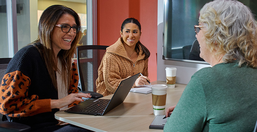 Three people are seated around a table in a casual office setting, smiling and engaged in a conversation. One person is working on a laptop, with coffee cups visible.