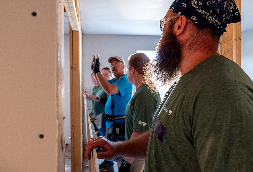 Three people in a partially completed room. One person holds a tool, pointing, while others watch attentively. Construction environment. Drywall installation likely.