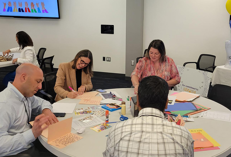 Three people are seated at a table with art supplies, actively engaged in what appears to be a crafting activity in a brightly lit room.