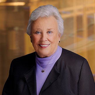 A professional portrait of a smiling person with short, gray hair, wearing a dark blazer, a purple turtleneck, and a necklace, set against a blurred background.