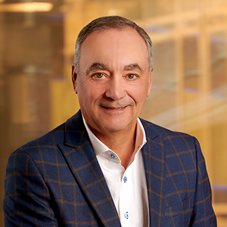 The image is a professional headshot of a smiling person wearing a patterned blazer over a white shirt, set against a blurred office background.