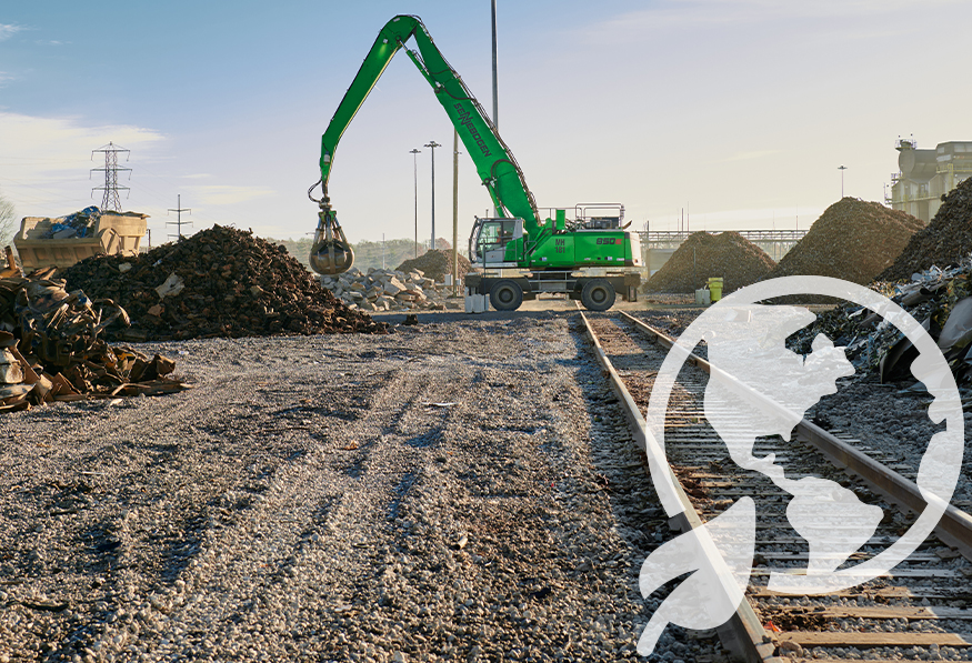 A green excavator with a grapple attachment stands amid piles of scrap metal and debris on industrial ground with train tracks and partial bridge visible.