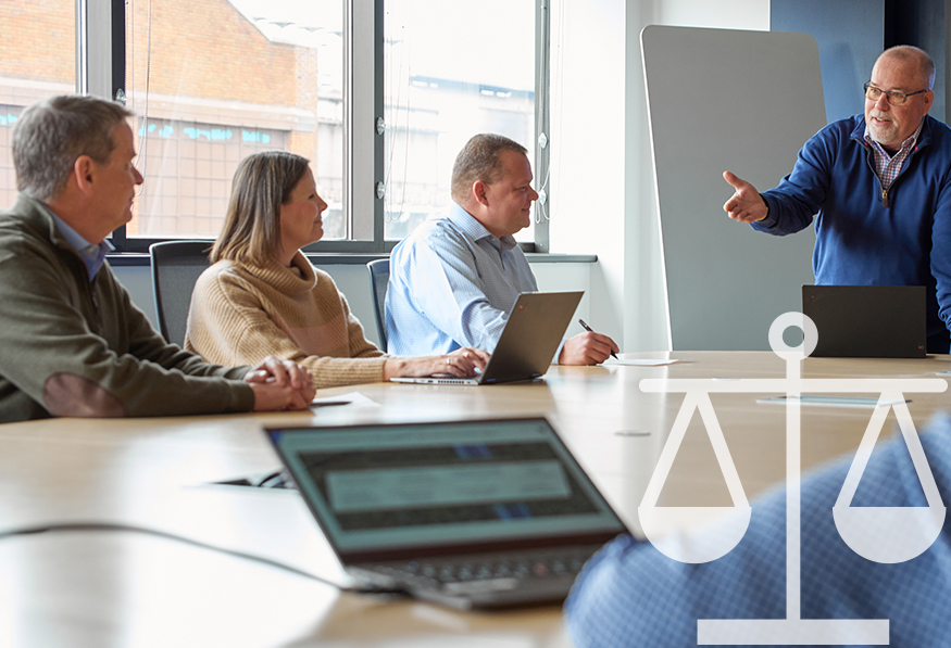 A group of people is in a meeting room with laptops. One person stands, presenting, while three others, seated at the table, listen attentively.