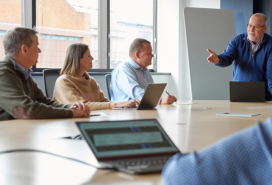 Three individuals sit at a conference table with laptops, attentively listening to a standing person presenting. The setting suggests a business meeting or workshop.