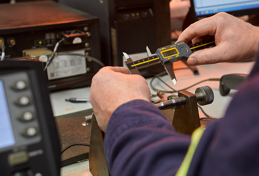 A person uses a digital caliper to measure an object on a bench surrounded by electronic equipment like an oscilloscope and vintage radio equipment.