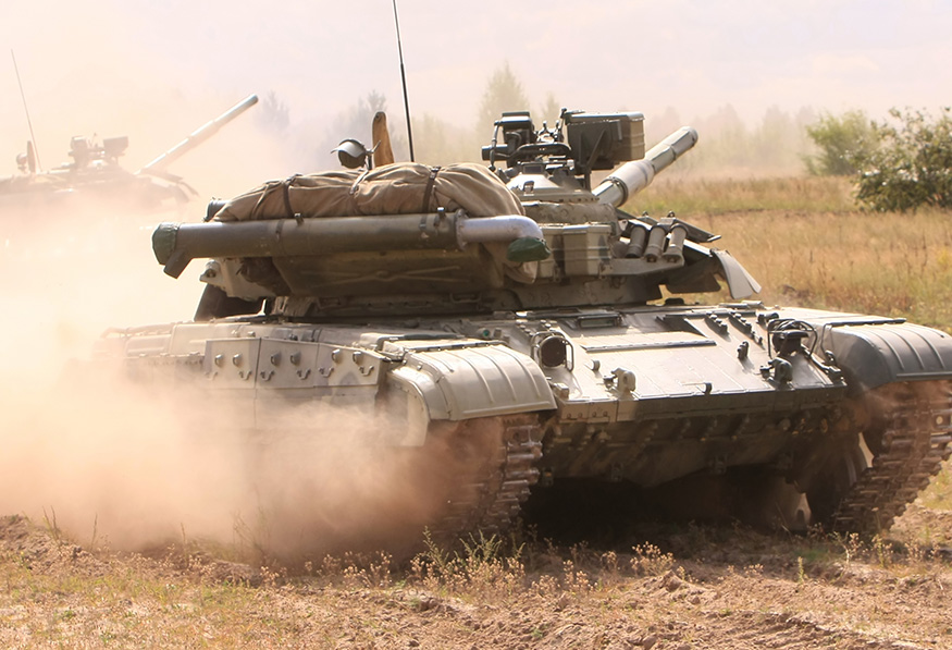 Two military tanks are advancing in a field, kicking up dust. Their main artillery cannons are prominent, and the scene suggests a training exercise or maneuver.