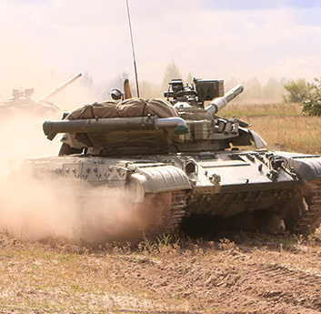 A military tank covered in dust advances through a dry field, its tracks churning the earth as it moves, with a turret and armaments visible.