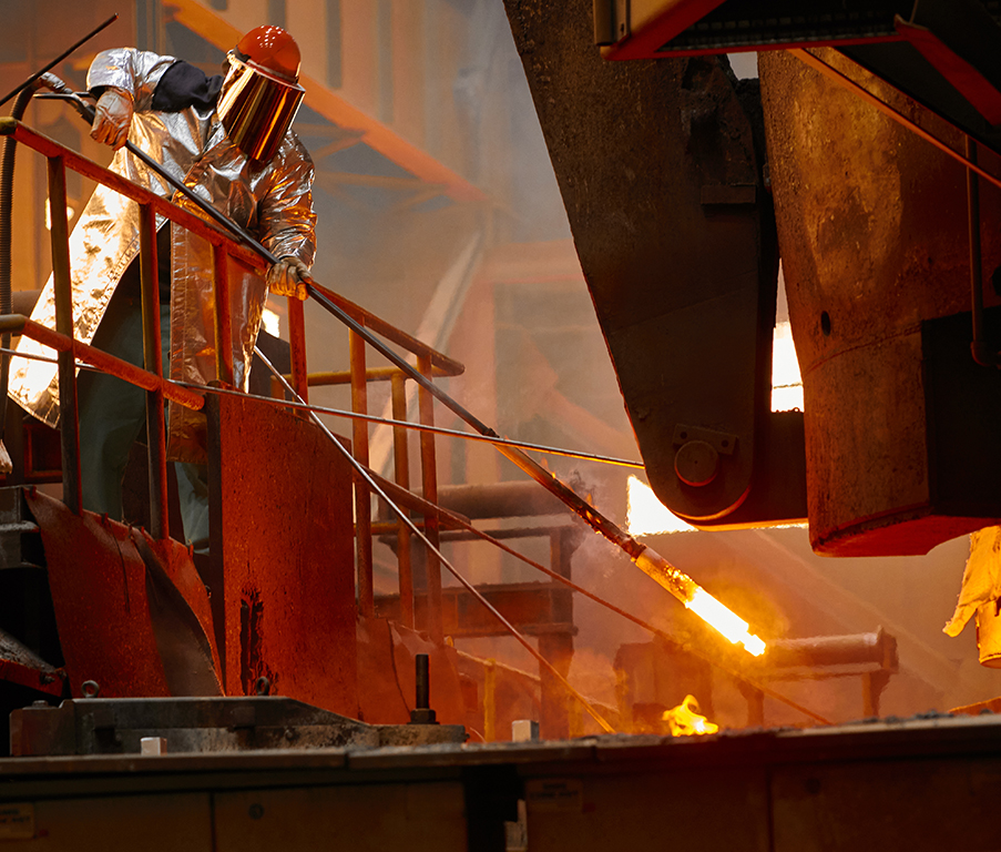 A person in protective gear is working with molten metal in an industrial setting, with intense heat and light visible around the machinery.