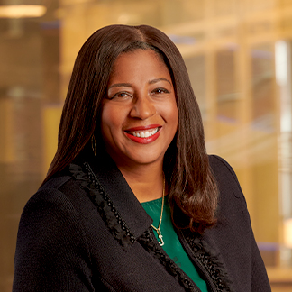 A professional portrait of a smiling person with long hair, wearing a black jacket, green top, and a necklace. Blurred office background suggests a corporate setting.