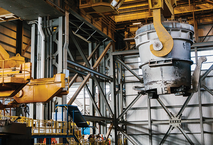 An industrial interior with yellow overhead cranes lifting a large metal ladle. Steel structures, machinery, and ductwork are visible in a factory setting.