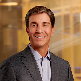 A person with short hair is smiling at the camera, wearing a business suit and shirt with no tie. They're in front of a blurred office background.