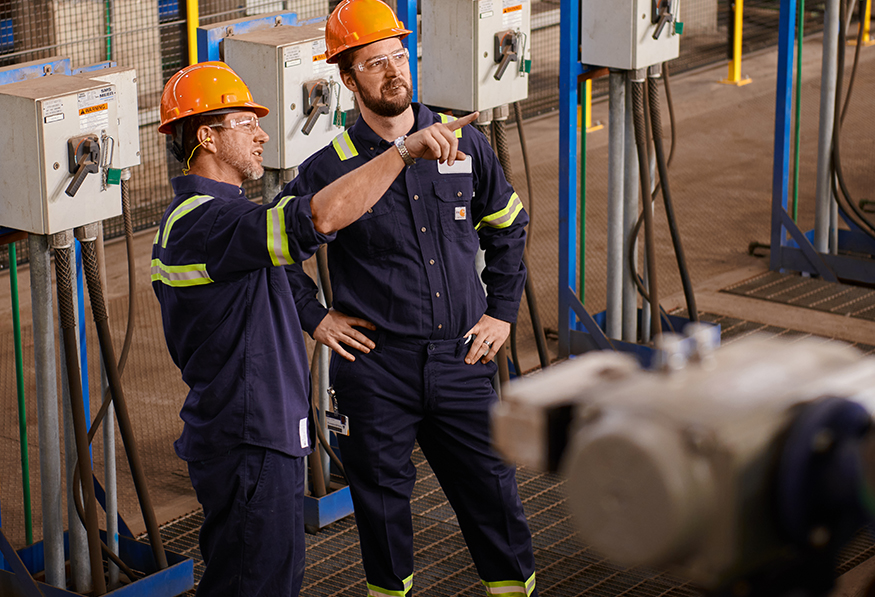 Two people wearing hard hats and reflective safety gear are in an industrial setting. One person is pointing and explaining something to the other.