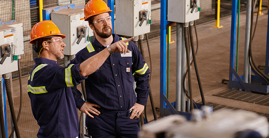 Two people in hard hats and high-visibility uniforms are discussing work in an industrial setting with machinery and control panels around them.