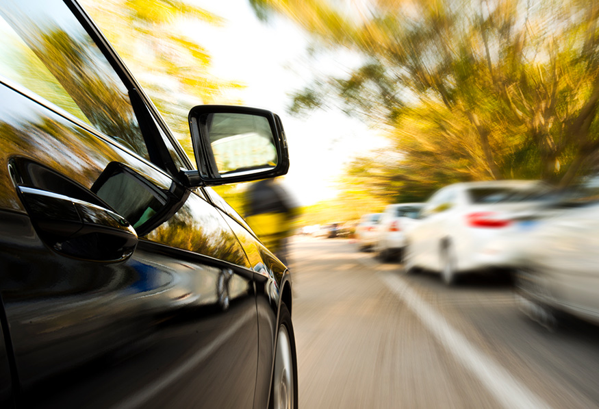 The image shows a motion blur effect from the side of a black car, with a focus on the rearview mirror, driving along a tree-lined road with other vehicles.