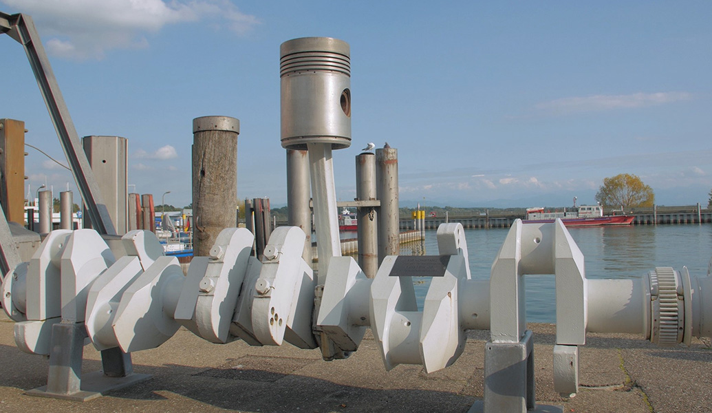 The image shows a large mechanical auger lying horizontally with a docking area and water in the background under a partly cloudy sky.