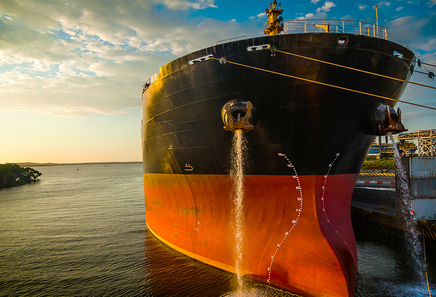 A large cargo ship with a rusty hull is moored at a dock. Water is being discharged from its ballast tanks at sunset.