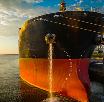 A large cargo ship with a black and red hull is docked, water draining from its ballast, against a backdrop of a vivid sunrise or sunset.