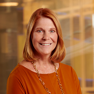 A smiling person with red hair, wearing an orange top and a gold necklace, is standing against a blurred office background.