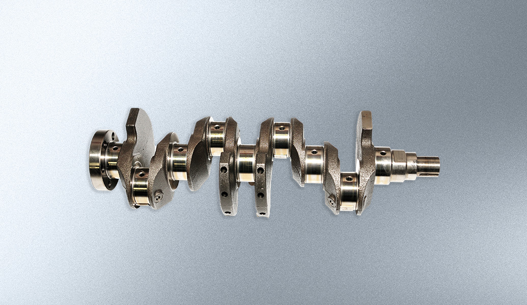 The image shows a metal car engine crankshaft with lobes and bearings against a grey background. It's an essential internal combustion engine component.