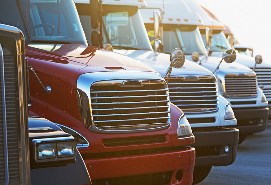 A lineup of semi-trailer trucks with chrome grilles parked in descending order, highlighted by warm sunlight, exuding a powerful, industrial atmosphere.