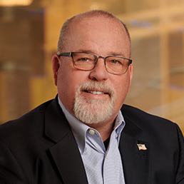 This image shows a professional headshot of a person with glasses, wearing a black suit and tie, with a flag pin on their lapel, smiling against a blurred background.