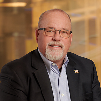 A person with glasses and a beard, wearing a black jacket, blue shirt, and a lapel pin, smiles in front of a blurred background.