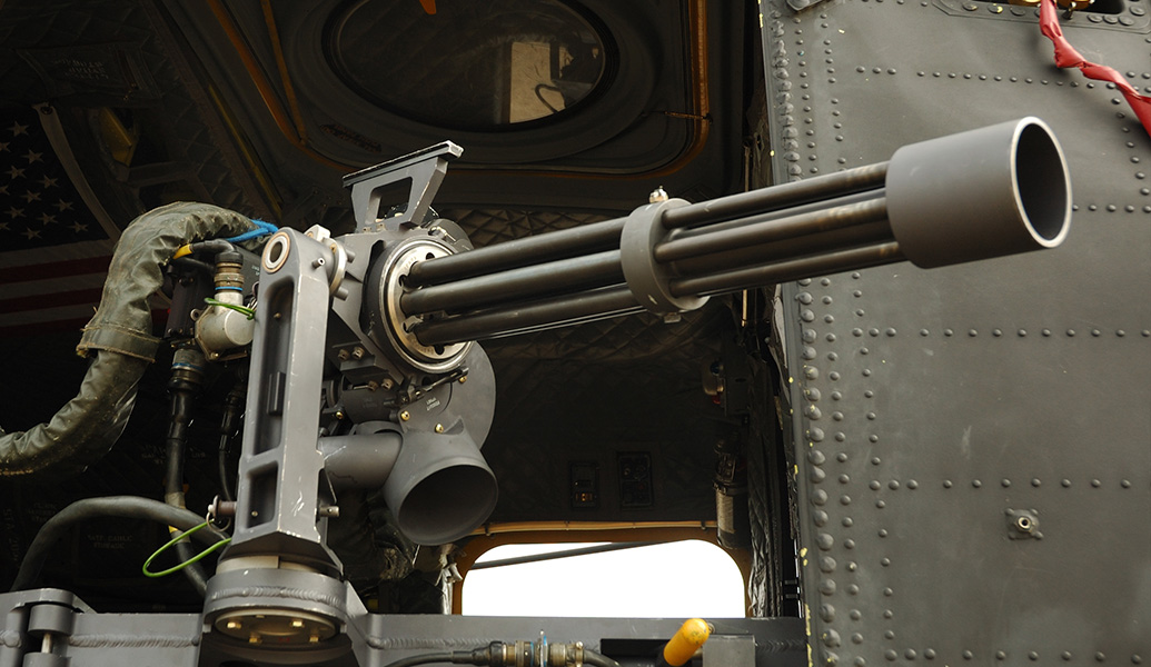 A person operates a mounted machine gun inside a military helicopter, with an American flag visible in the background, indicating a likely U.S. setting.