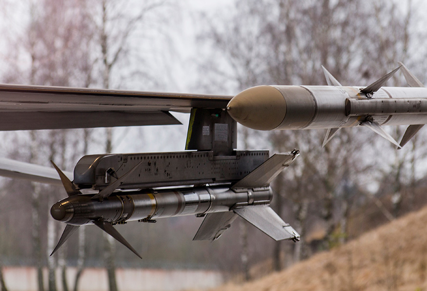 The image shows a close-up of a military aircraft's wing with mounted missiles and precision-guided munitions, against a blurred natural background.