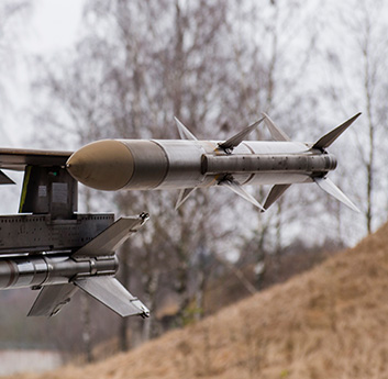 Close-up of a missile attached to the underside of an aircraft wing, with a blurred natural background of trees and a grassy slope.