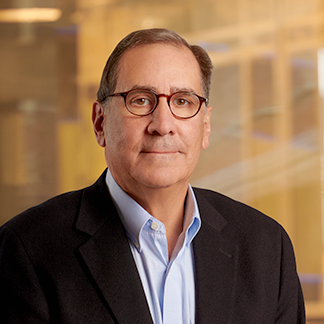 A person is pictured with glasses, wearing a dark blazer over a blue shirt, against a blurred indoor background, possibly an office setting.