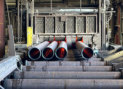 Four large red-tipped pipes are lying on a conveyor system inside an industrial facility. Chains hang above, suggesting a loading or manufacturing process.