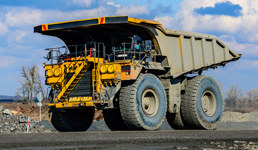 A large yellow mining dump truck is shown with a massive load capacity, featuring huge wheels, driving on a rocky surface with a clear sky above.