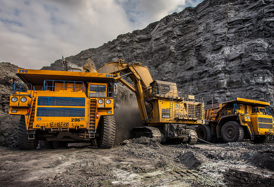 Two large yellow mining trucks are being loaded with material by a massive excavator in an open pit mine with exposed dark rock faces.