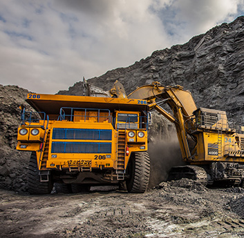 A large yellow mining dump truck is being loaded with dirt by an excavator in an open-pit mine, with a cloudy sky above and rocky terrain.