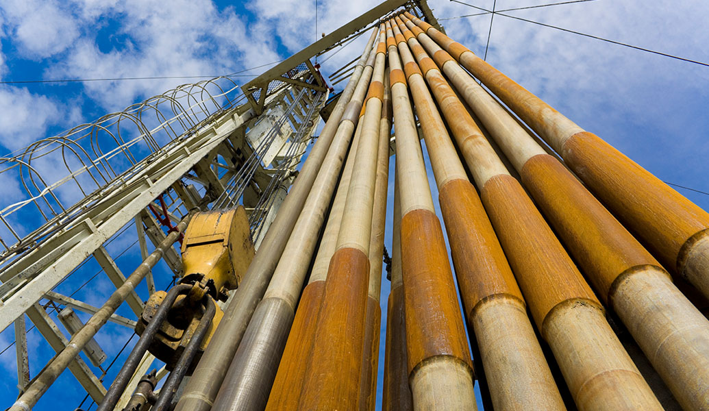 The image shows a low-angle view of long, rusted industrial pipes extending upwards against a blue sky with clouds, possibly part of an oil rig or factory.