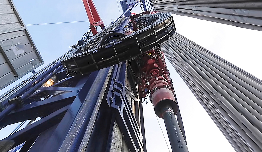 The image shows a close-up view of an industrial drilling rig machinery with cables and hydraulic mechanisms, against a clear sky. No people are visible.