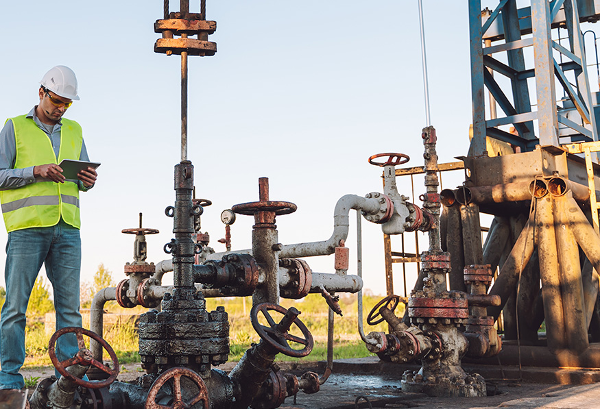 A person in a safety helmet and high-visibility vest inspects an industrial oil pump site with complex piping, using a digital tablet during golden hour.