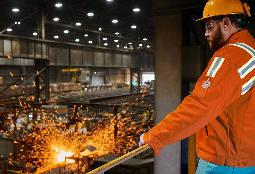 A person in safety gear works in an industrial setting with molten materials, sparks flying. The environment looks busy, focused on metal fabrication or smelting.