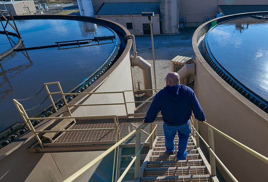 A person descends a metal staircase between two large, circular water treatment tanks under a clear blue sky, with industrial buildings and water in the background.