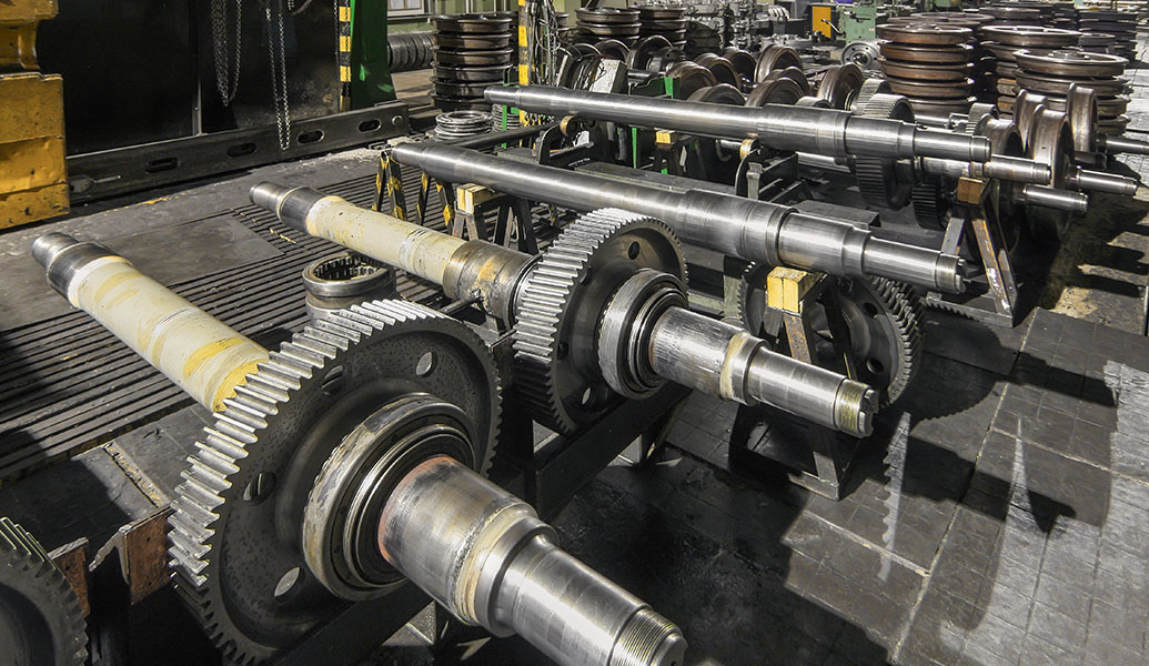 The image displays an industrial setting with several metal shafts and gears, possibly part of machinery or vehicle components, laid out on a factory floor.