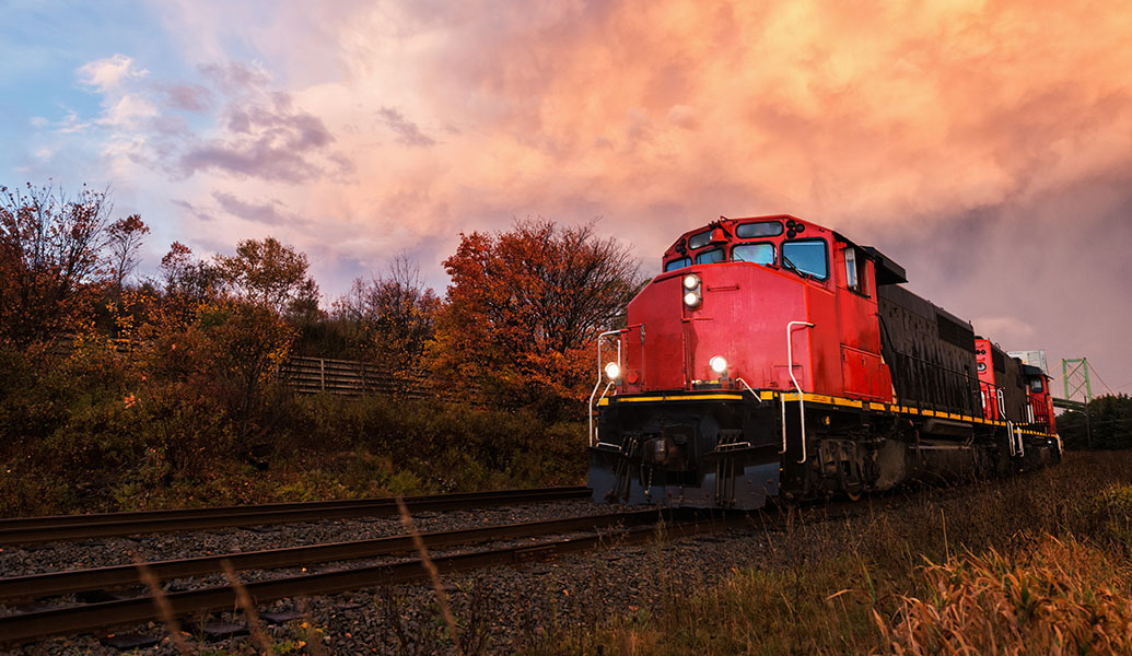 A red freight train moves along tracks amidst autumn scenery under a dramatic sky illuminated by the warm glow of the sunset.