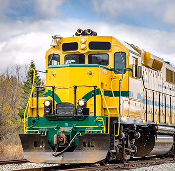 A bright yellow and green locomotive on train tracks, with a cloudy sky backdrop, no visible people, and early signs of fall in the surrounding foliage.