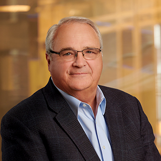 A person with glasses wearing a blazer over a light blue shirt, smiling, with a blurred office background. Professional headshot, possibly for corporate use.