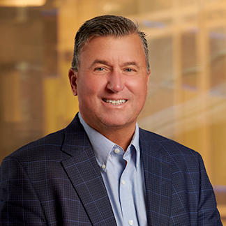 This is a professional portrait of a smiling person wearing a blue checkered suit and light blue shirt, set against a blurry indoor background.