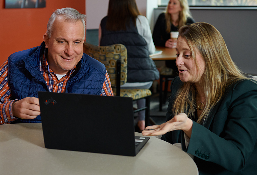 Two people are sharing a moment at a table with a laptop. The person on the right is gesturing animatedly, possibly explaining something to the smiling person.