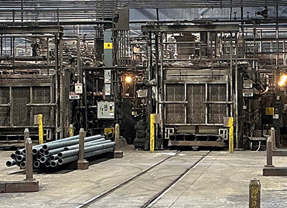 The image shows an industrial setting with machinery, pipes on the floor, rail tracks, and a forklift. It has a gritty, functional appearance, typical of manufacturing facilities.